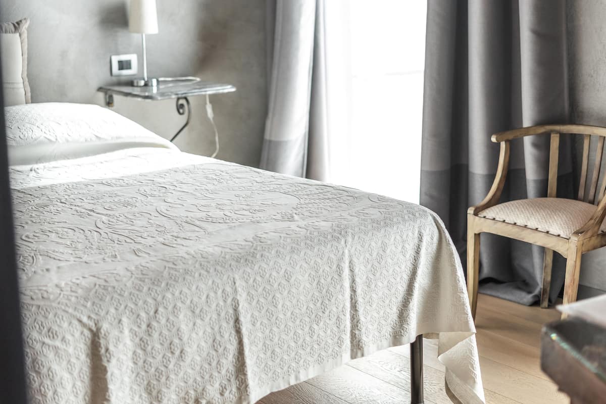 Un lit bien fait, recouvert d’une couvre-lit à motifs, une chaise en bois avec un coussin, et une table de chevet avec une lampe et un petit appareil.