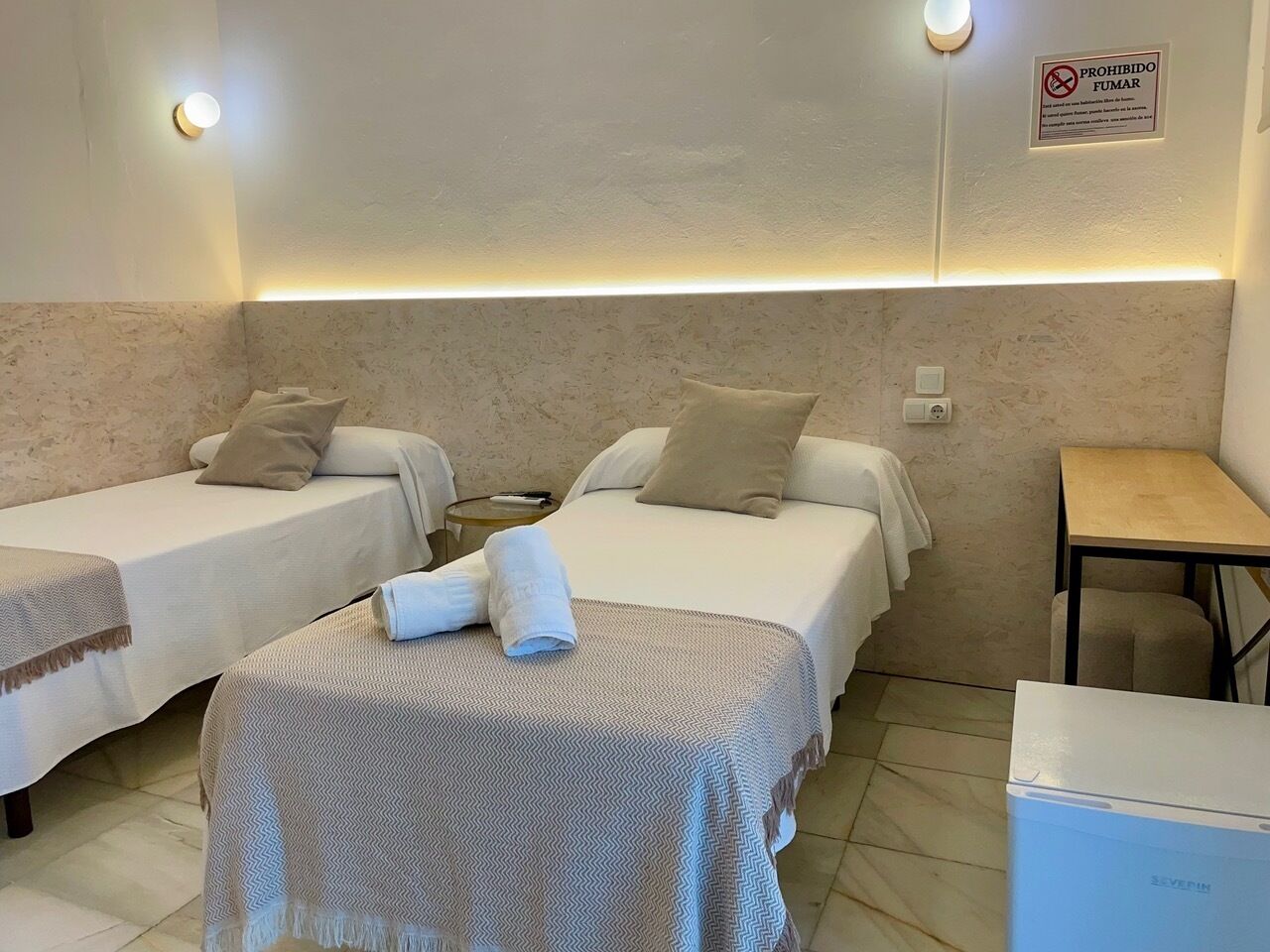 A hotel room with two beds, a small table, a chair, and a wall-mounted light fixture.