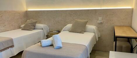A hotel room with two beds, a small table, a chair, and a wall-mounted light fixture.