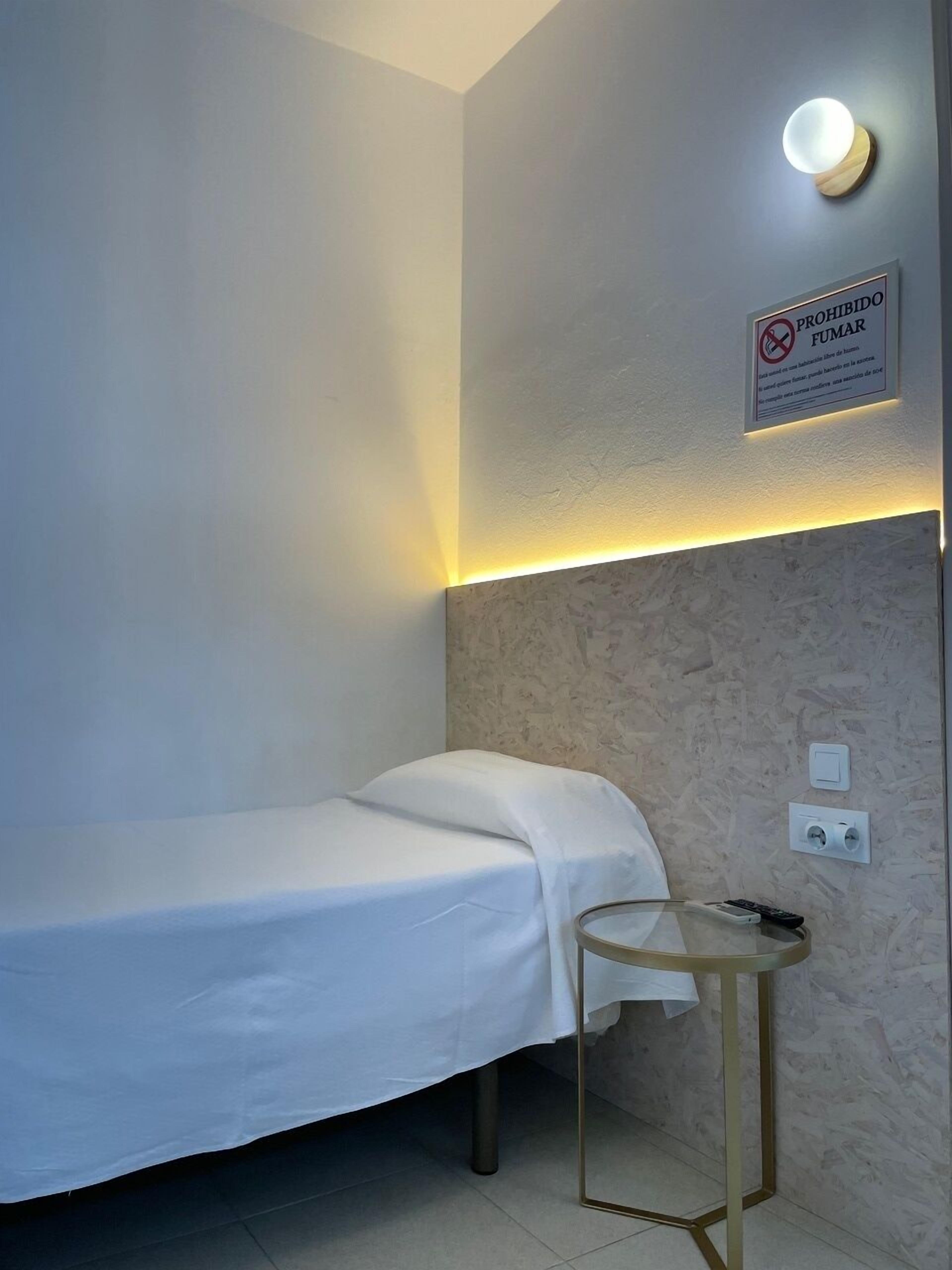 A single bed with a white sheet, a small bedside table, a wall-mounted light, and a sign prohibiting smoking.