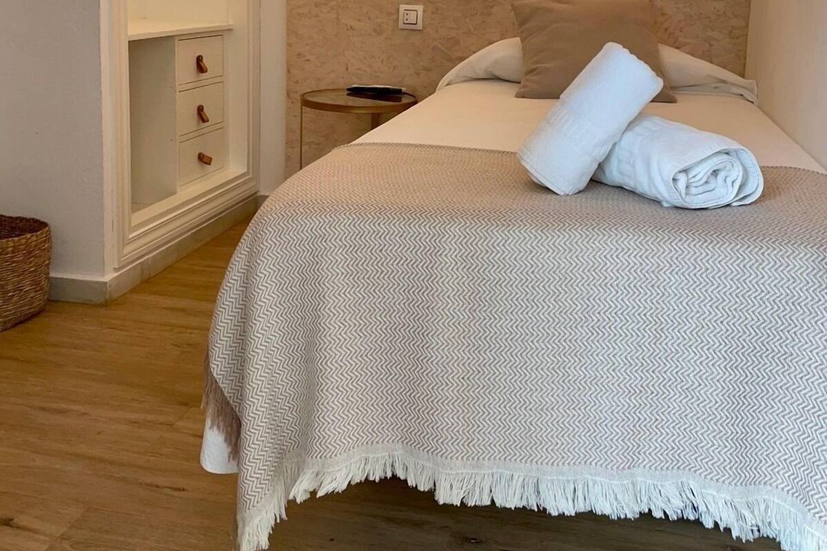 Een net opgemaakt bed met een witte deken en een opgerolde handdoek, een nachtkastje met een afstandsbediening, en een wandlamp.