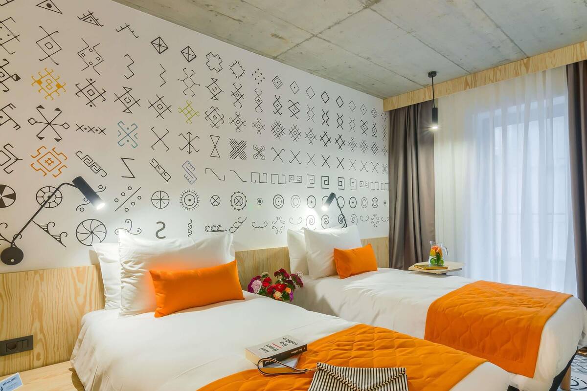 A hotel room with two beds, a wall with geometric patterns, a bedside table with a lamp, and a window with curtains.