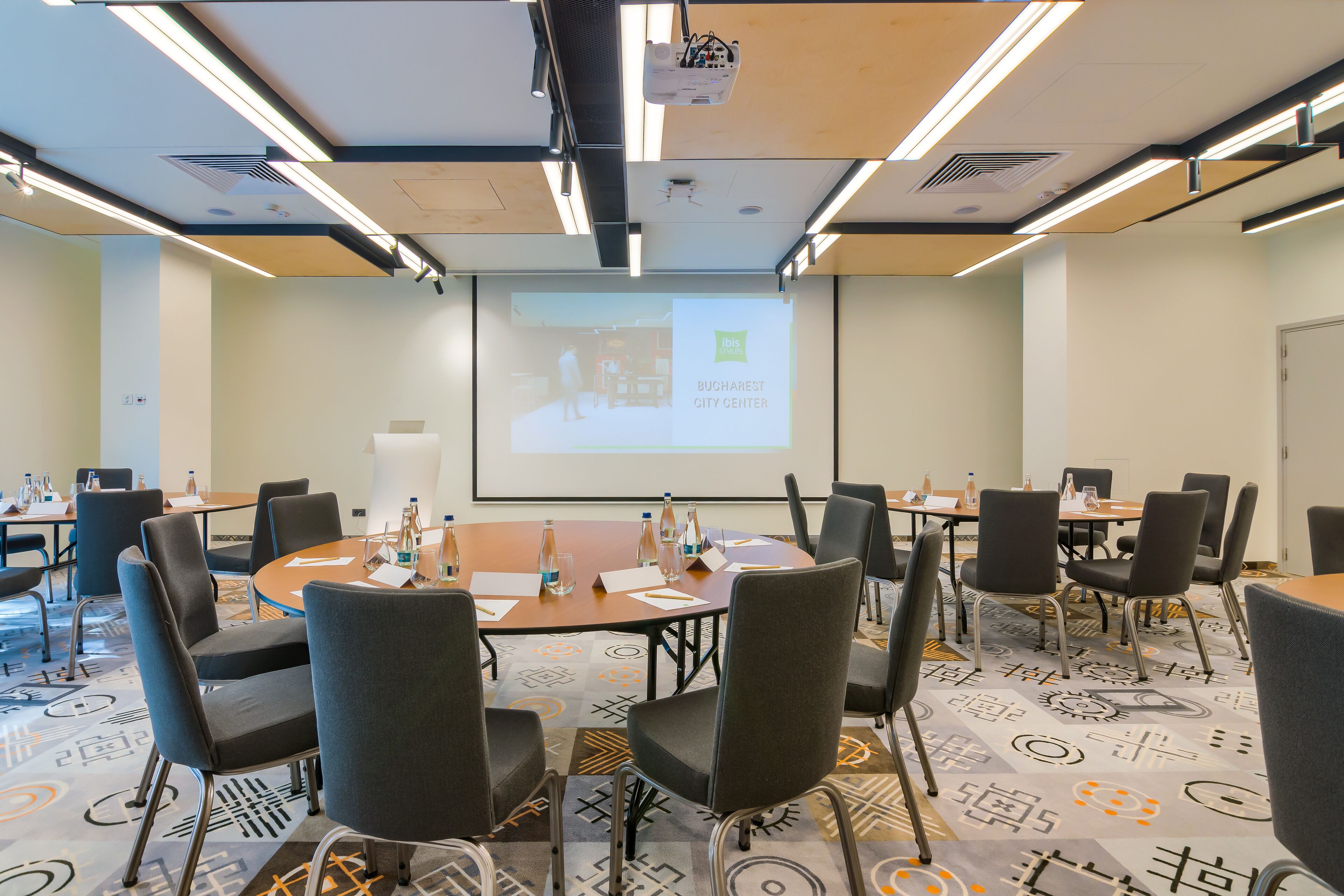 A conference room with a large screen displaying a presentation, multiple tables with chairs, and water bottles on the tables.