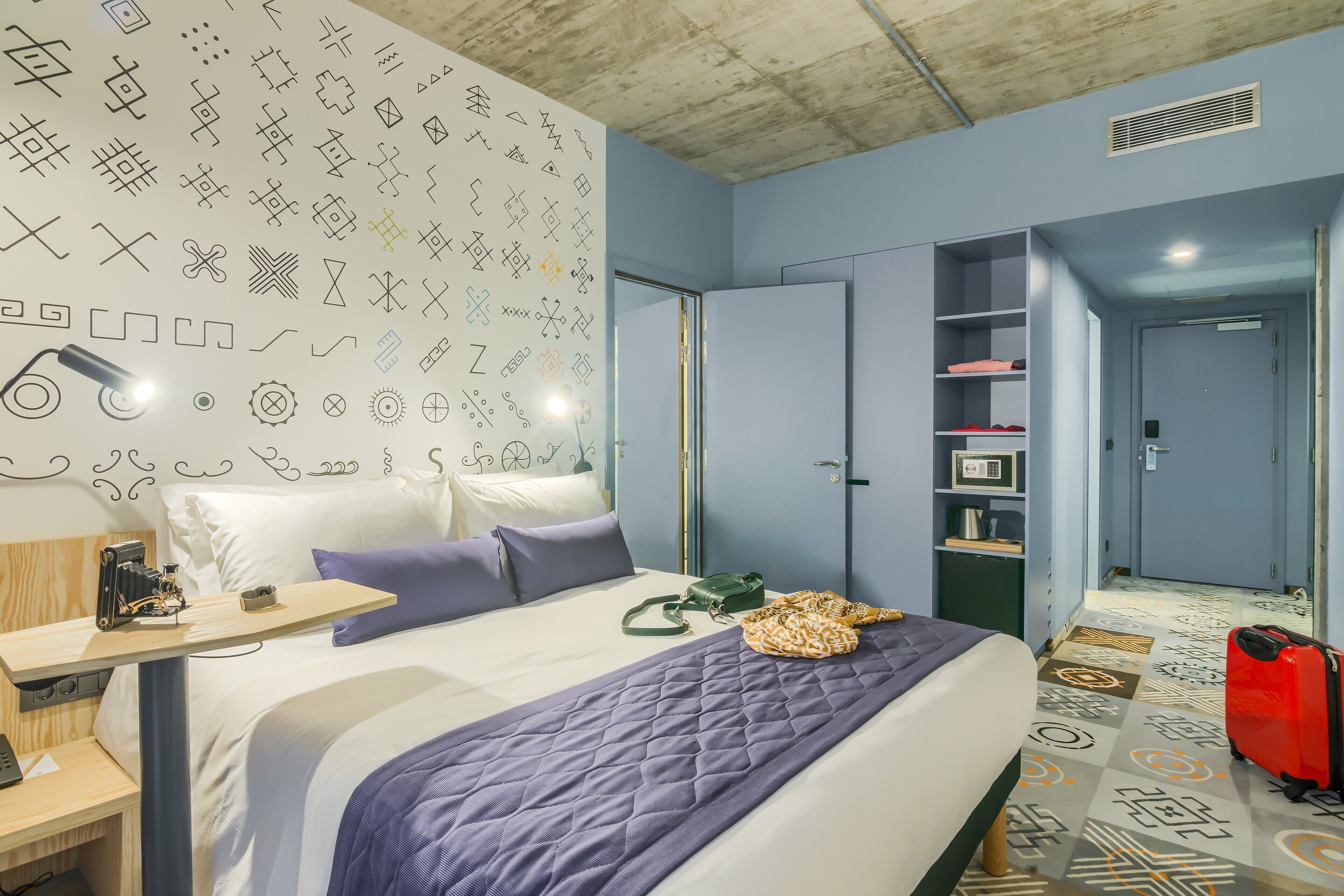A hotel room with a bed, a desk, a wardrobe, and a patterned floor.