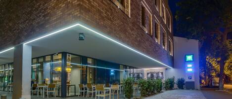 An outdoor cafe area with tables and chairs, a brick building facade, and a digital sign displaying the time.
