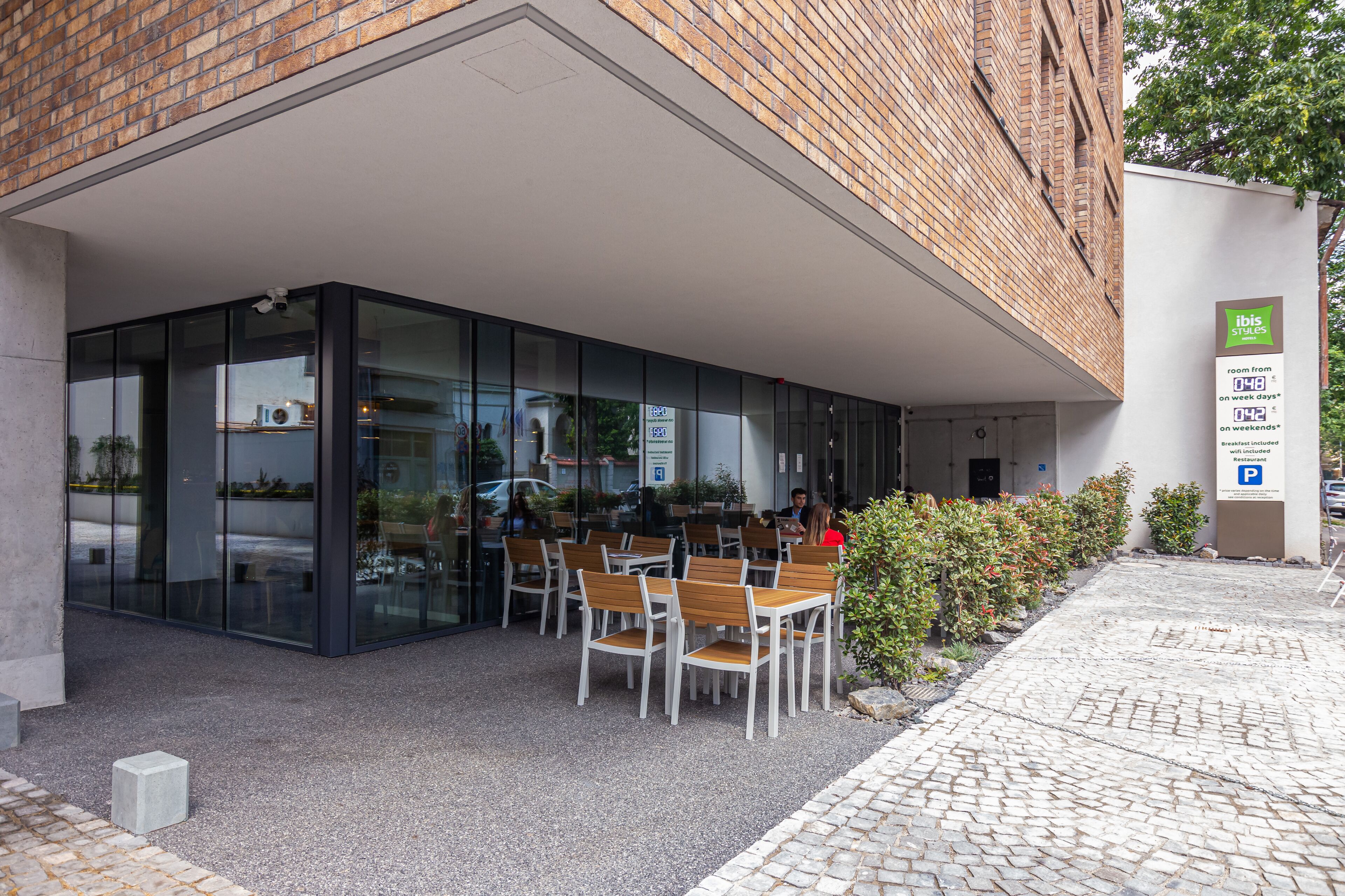 Un espacio al aire libre con mesas y sillas, un muro de cristal y un cartel que indica las tarifas de las habitaciones.