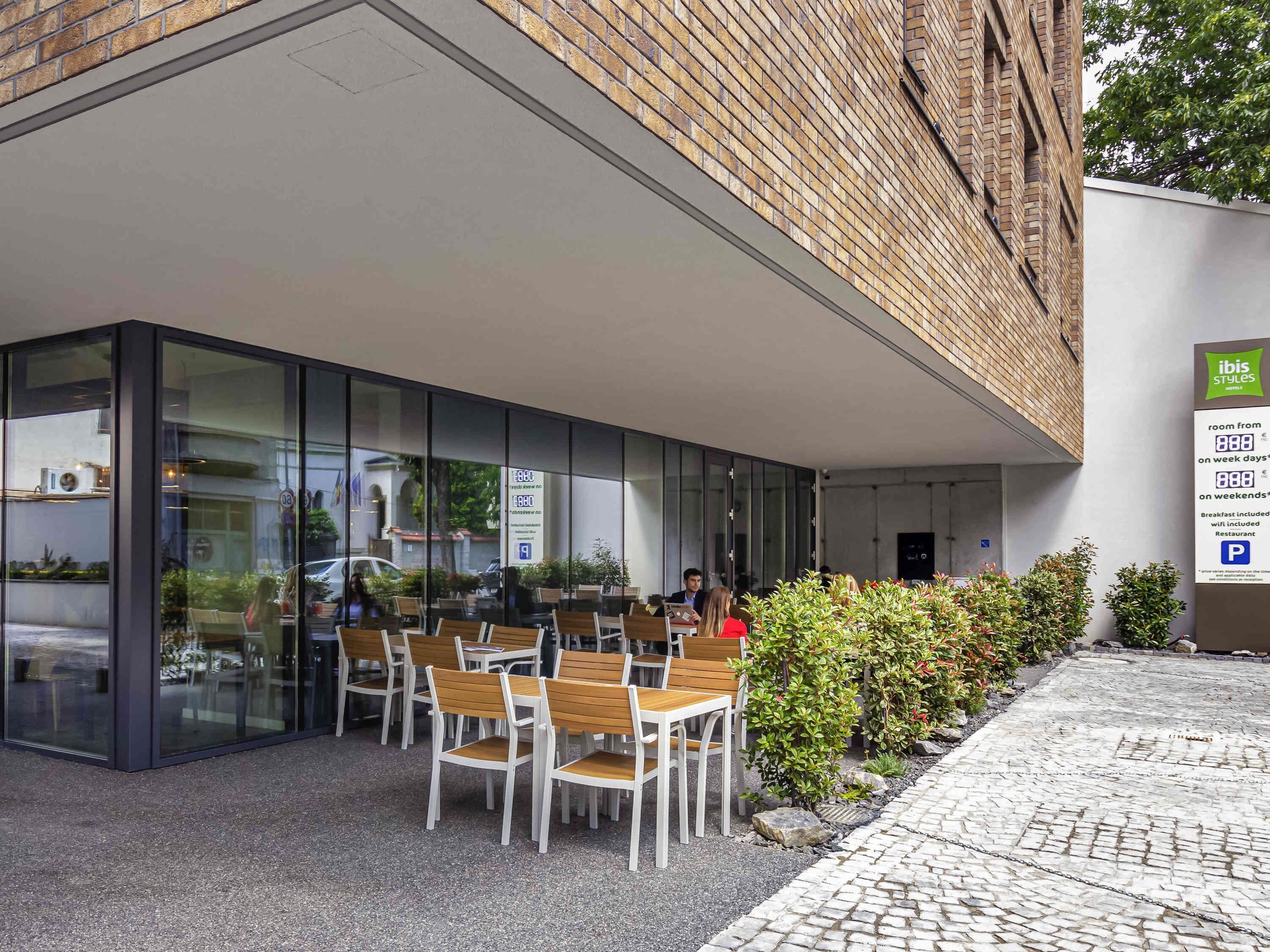 Een hotelentree met terras, een glazen gevel en een bord met prijzen voor de kamers.