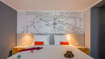 Habitación de hotel con un gran mapa mural, una cama con ropa blanca, dos almohadas, un plaid rojo, un sombrero y un libro.