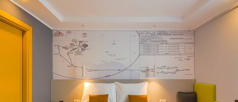 A hotel room with a large wall map, a bed with white bedding, two pillows, a bedside table with a lamp, and a bench with a cushion.