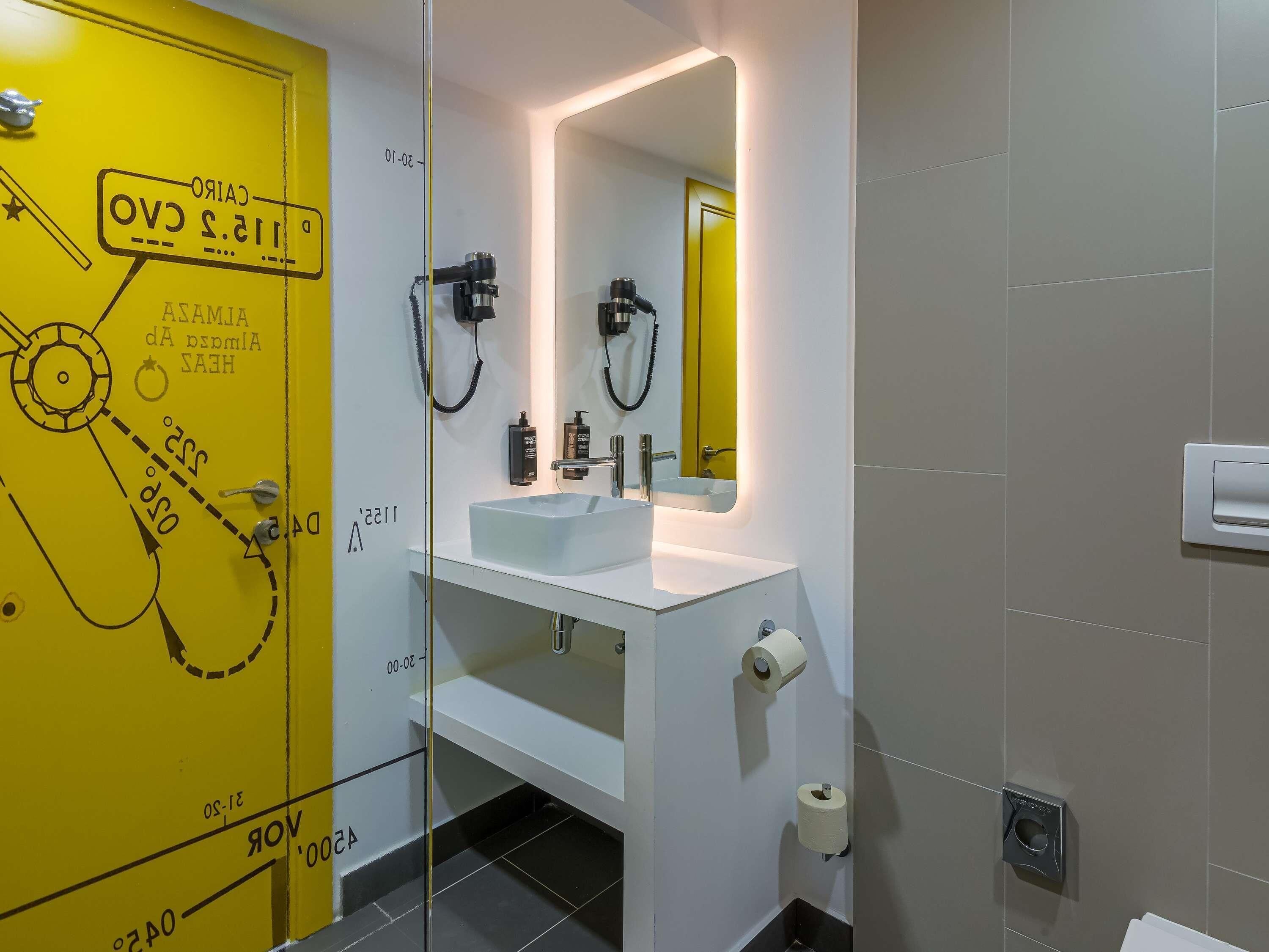 Un baño moderno con una puerta amarilla, un lavabo blanco, un espejo y un dispensador de papel higiénico.