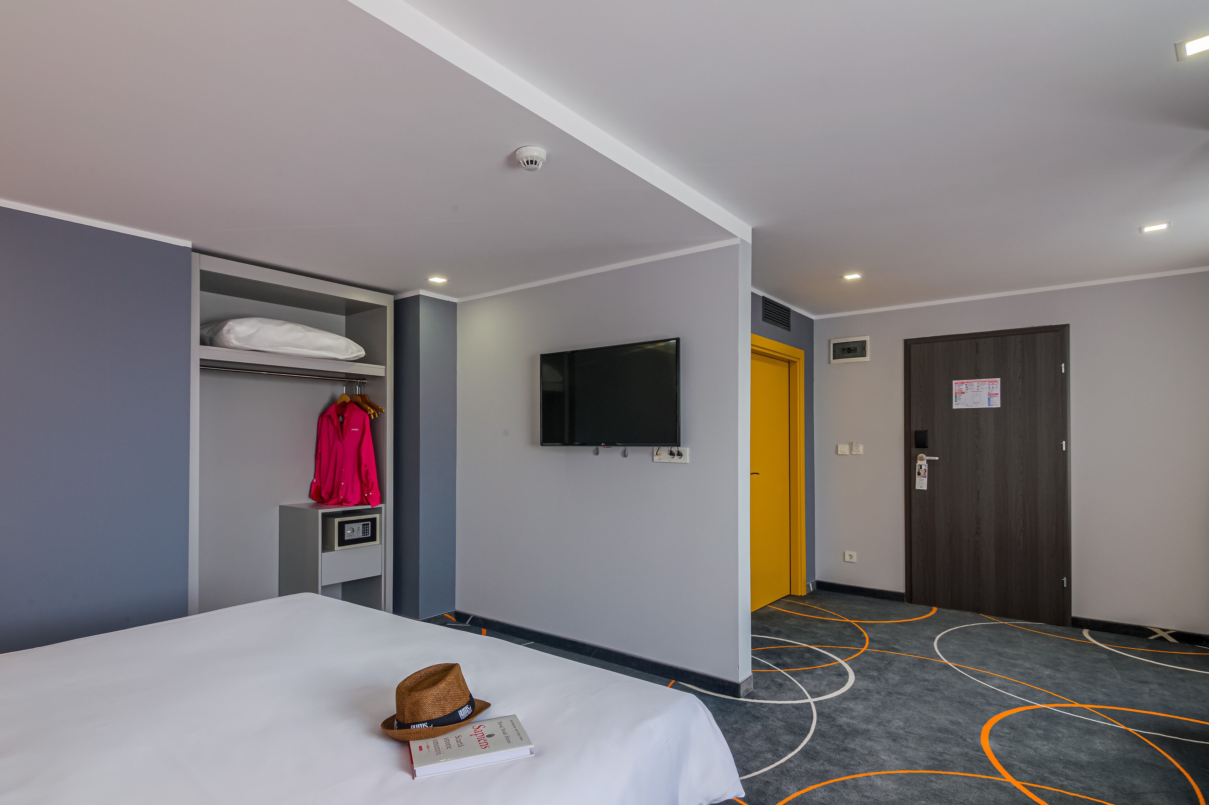 Una habitación de hotel con cama, televisor montado en la pared, armario, perchero y puerta.