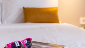 Un lit avec des oreillers blancs et moutarde, une paire de lunettes de soleil roses posée sur un magazine et une table de chevet avec une lampe.