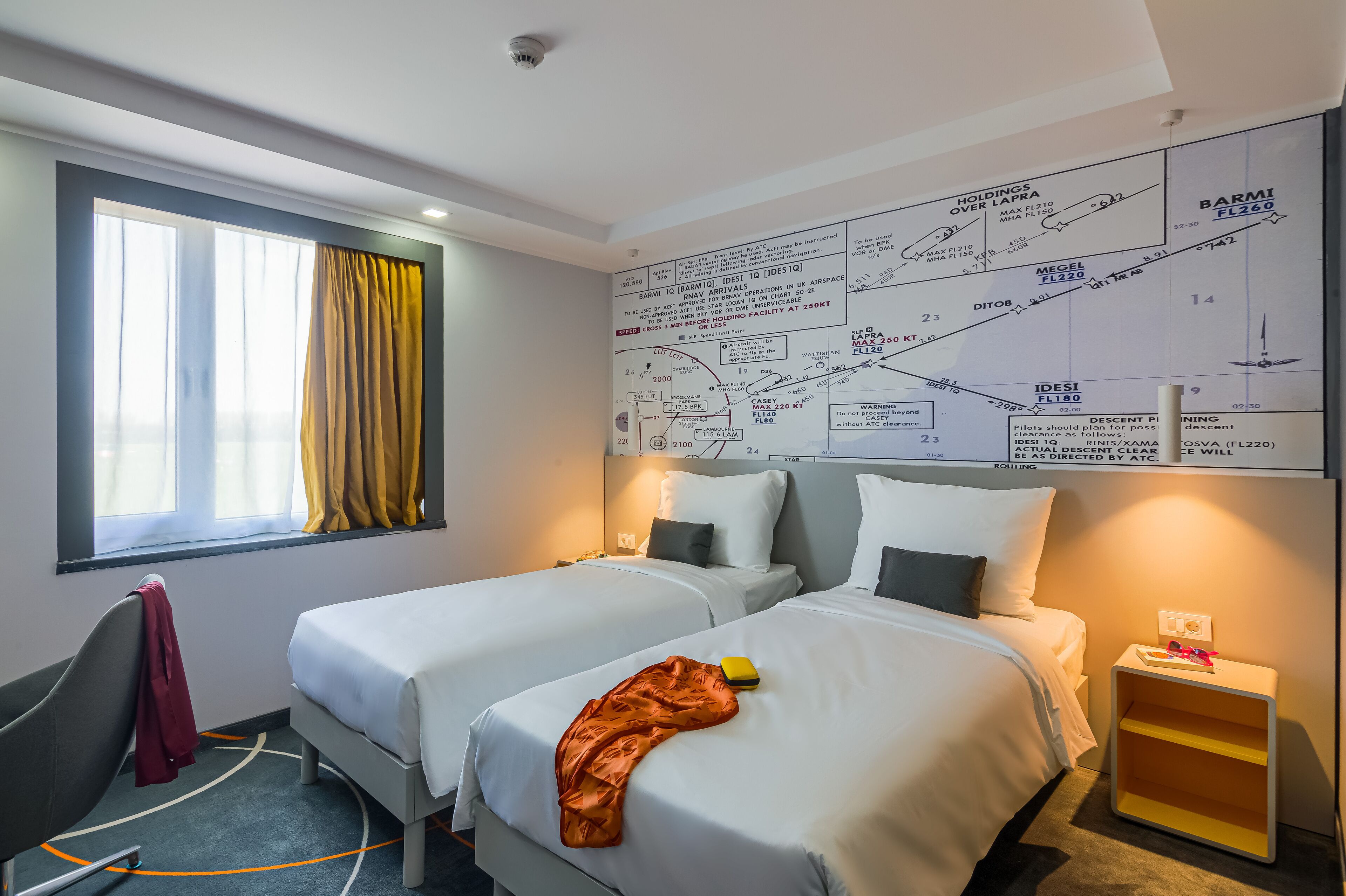 Habitación de hotel con dos camas, una ventana con cortinas amarillas, un mapa en la pared y una mesita de noche pequeña con una lámpara.