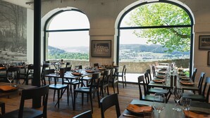 Een restaurant met gedekte tafels, grote gewelfde ramen met uitzicht op een vallei, en een ingelijste historische foto aan de muur.