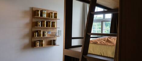 Una habitación con una estantería de madera que muestra varios recipientes, una ventana con vista a follaje y una cama con una colcha estampada.