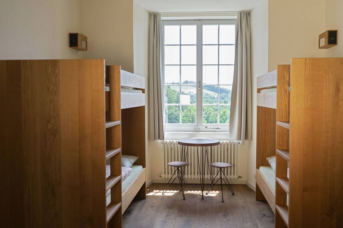 Una habitación con literas de madera, una mesita y una ventana que da a zonas verdes.
