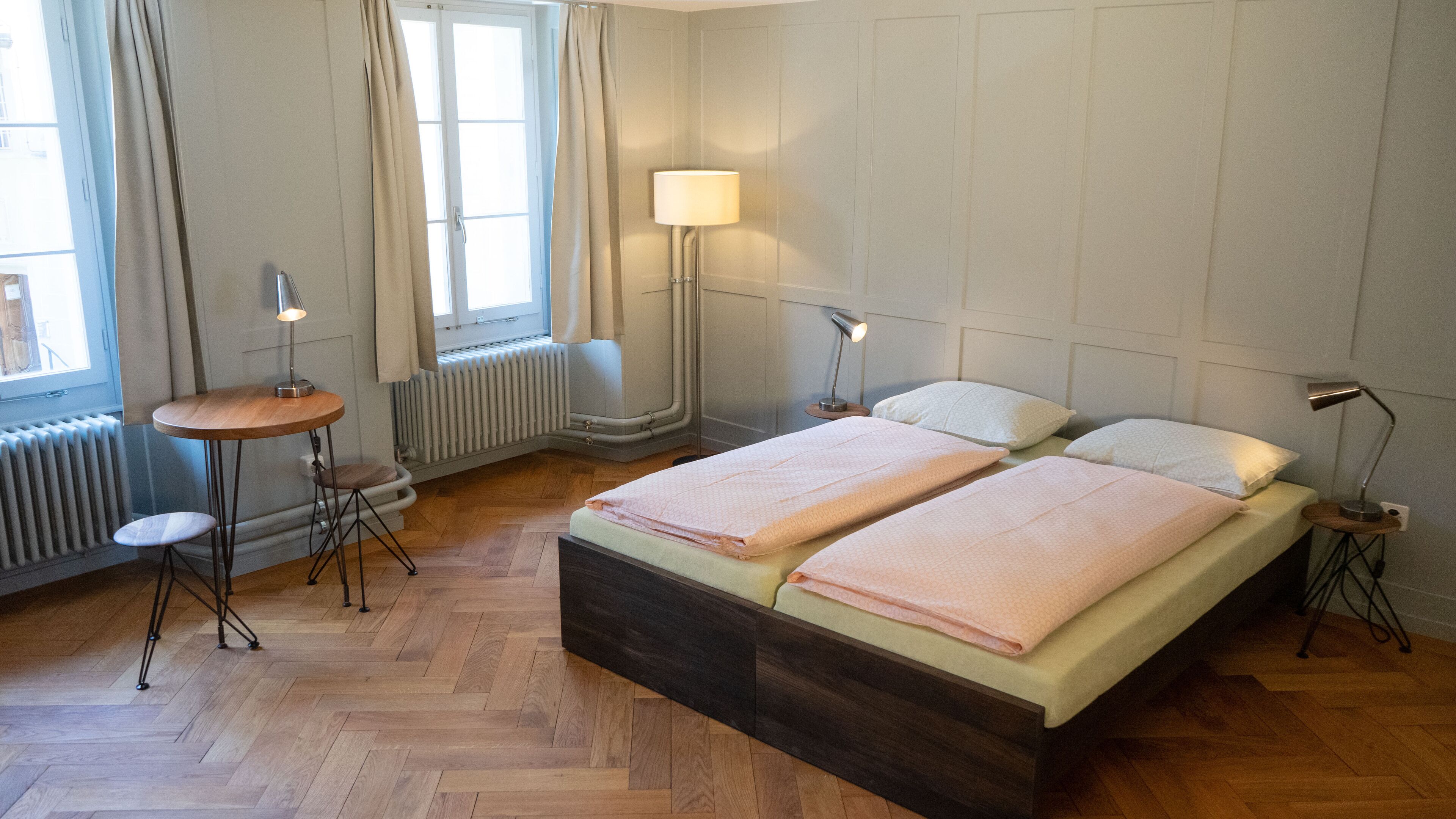 A hotel room with a double bed, two bedside lamps, a small round table with a chair, a radiator, and a window with curtains.