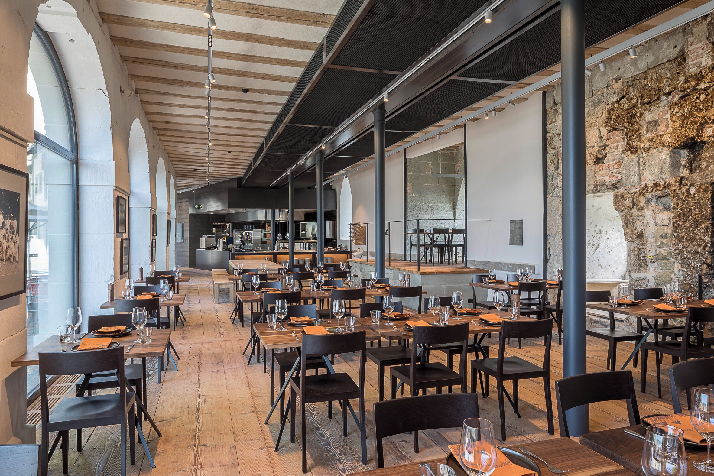 A spacious restaurant with high ceilings, wooden beams, and large windows. Tables are set with wine glasses and plates.