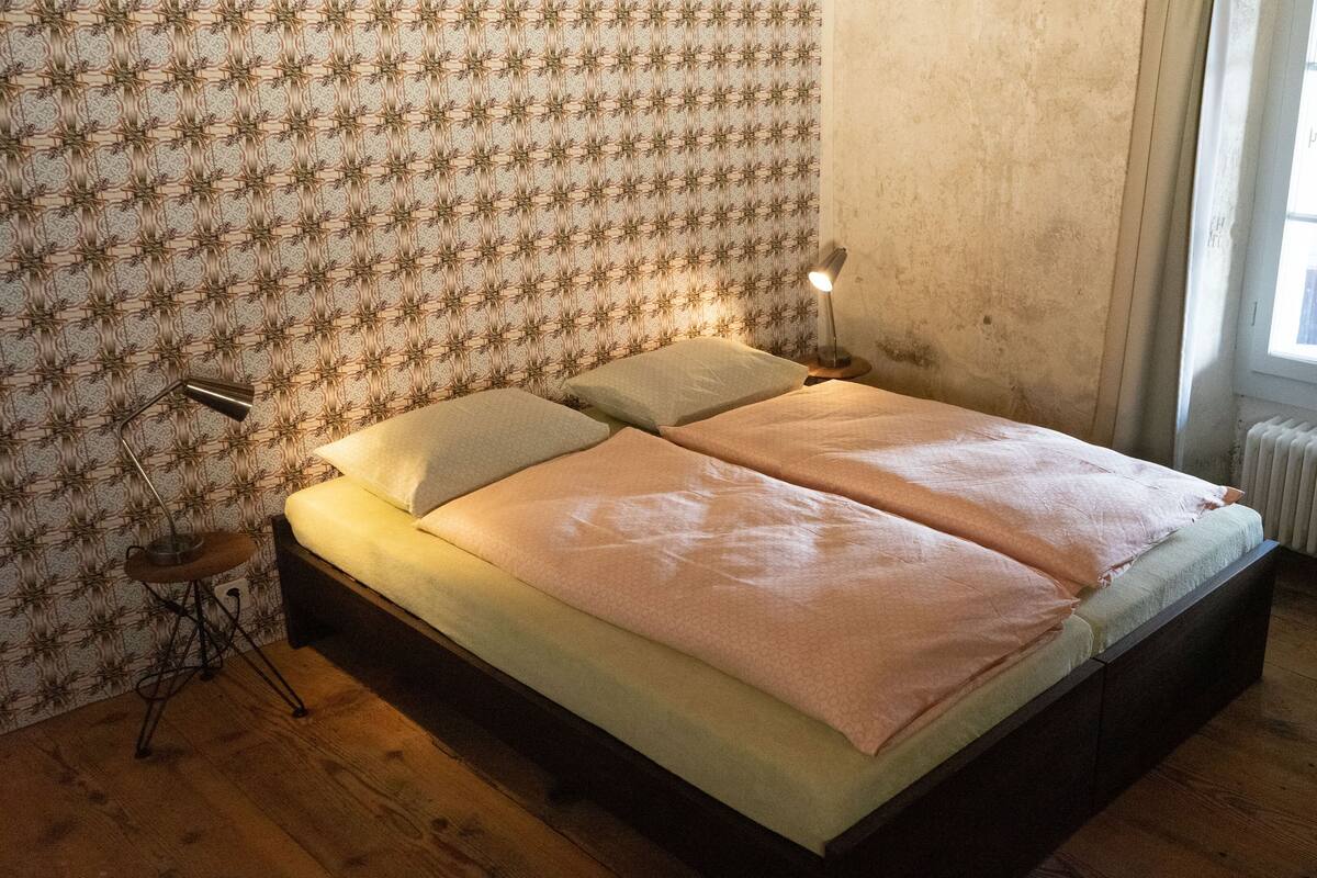 Un dormitorio con suelo de madera, papel tapiz estampado, cama con dos almohadas, lámpara de noche y radiador.