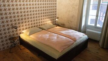A bedroom with a wooden floor, a patterned wallpaper, a bed with two pillows, a bedside lamp, and a radiator.