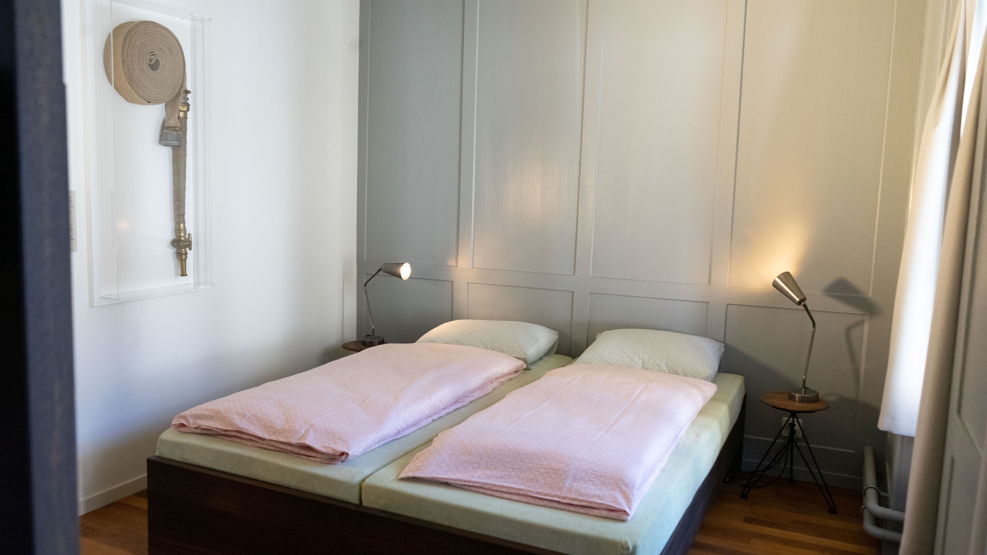 Un lit double avec deux oreillers, une table de chevet, une lampe et une lance d’incendie fixée au mur.