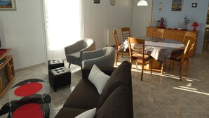 A living room with a brown sofa, a dining table with chairs, a TV, and a window with curtains. - Charming F3 house in Saujon, 2 bedrooms, pets allowed, parking (Saujon)
