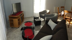 A living room with a TV on a wooden stand, a sofa, a chair, a small table with red stools, and a dining table with chairs. - Charming F3 house in Saujon, 2 bedrooms, pets allowed, parking (Saujon)