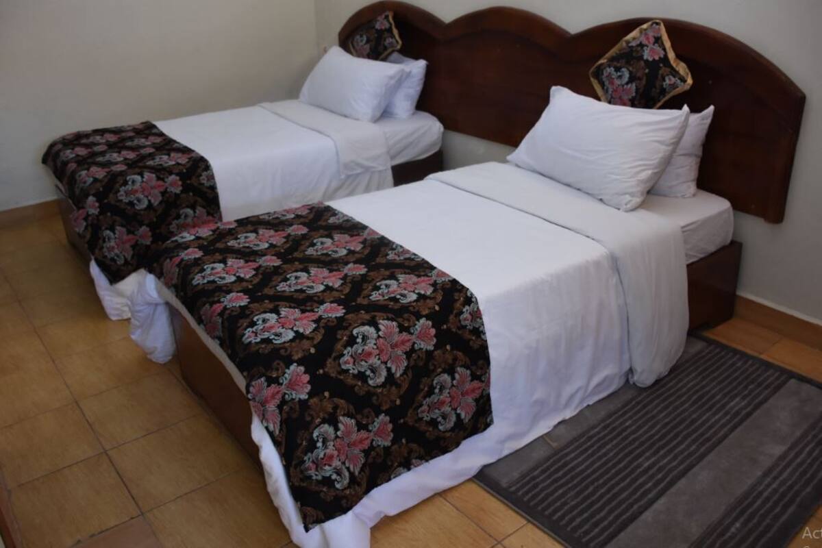 A room with two beds, each with a wooden headboard and white bedding. There is a dark-colored rug in front of the beds.