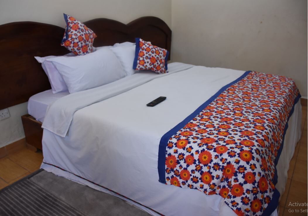 Una cama bien hecha con sábanas blancas y una manta estampada con motivos florales, un control remoto sobre la cama y un cabecero de madera.