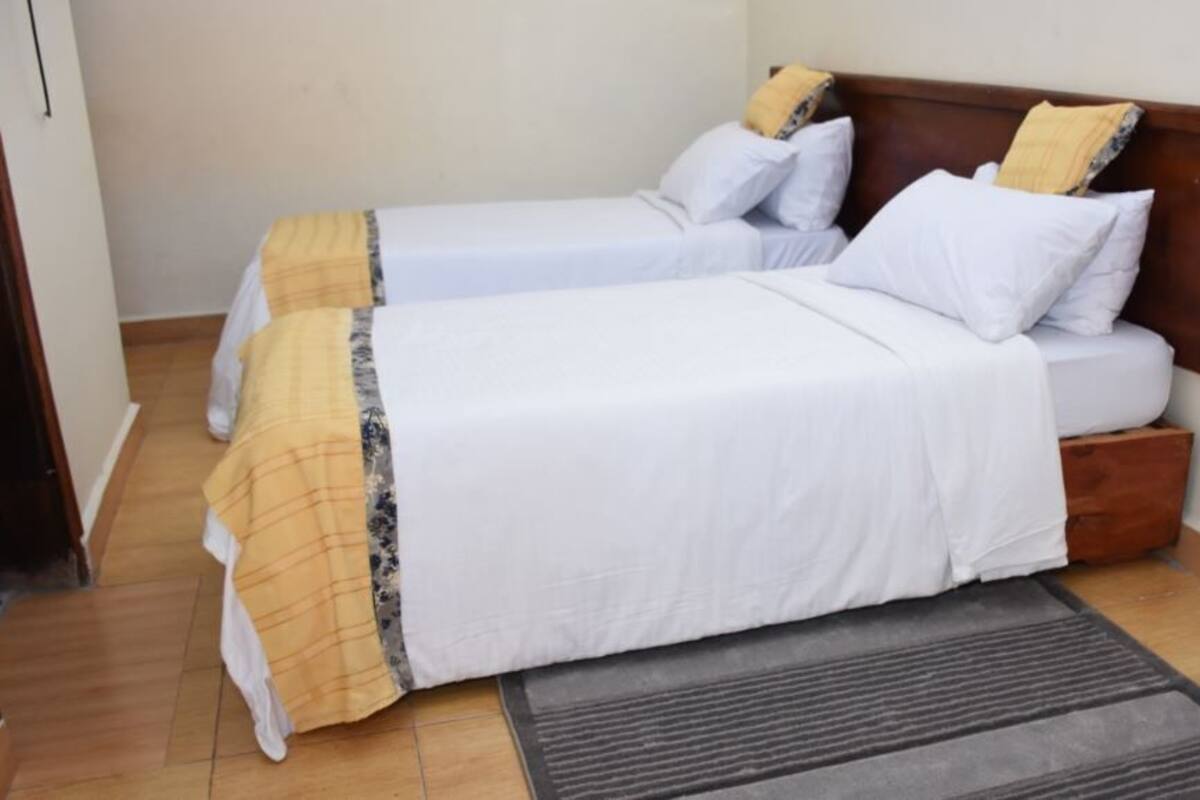 A hotel room with two single beds, a wooden headboard, a small wooden nightstand, and a grey rug on the floor.