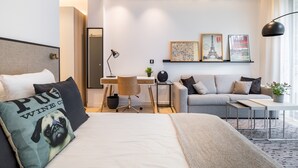 Ein modernes Hotelzimmer mit einem Bett, einem Sofa, einem Schreibtisch und einem kleinen Tisch. An der Wand hängen gerahmte Bilder und auf dem Tisch steht eine Pflanze.