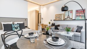 Ein modernes Hotelzimmer mit Essbereich, einer Couch und einem Couchtisch. An der Wand hängen Bilderrahmen, eine Lampe steht auf dem Couchtisch und eine Pflanze ebenfalls.