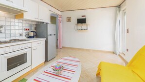 Cocina compacta con estufa, refrigerador y microondas. Hay una mesita con un objeto de colores encima. Se ve una silla amarilla en la esquina.