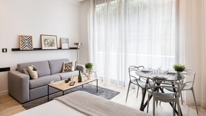 A modern living room with a grey sofa, a coffee table, and a dining table set for four. There are framed pictures on the wall, a floor lamp, and a large window with sheer curtains. - A3 Suite Studio 175Blo-Paris Convention (Paris-15E-Arrondissement)