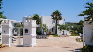 A gated entrance to a modern building with a paved driveway, palm trees, and bicycles parked outside. - Torre Guaceto Greenblu Resort (Carovigno)