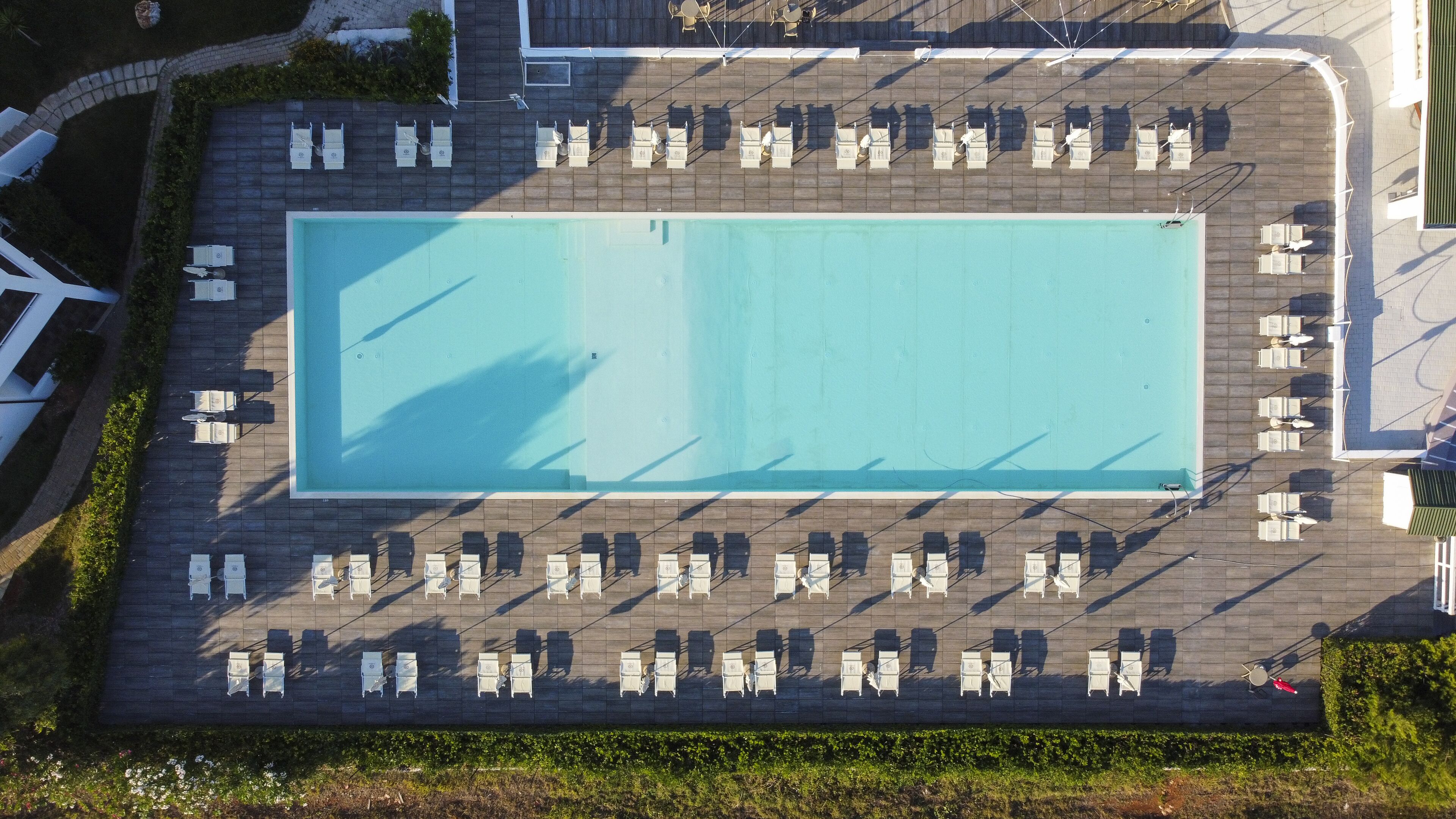 Une vue aérienne d’une piscine rectangulaire entourée de rangées de chaises longues et de fauteuils, avec un chemin pavé et de la végétation tout autour.