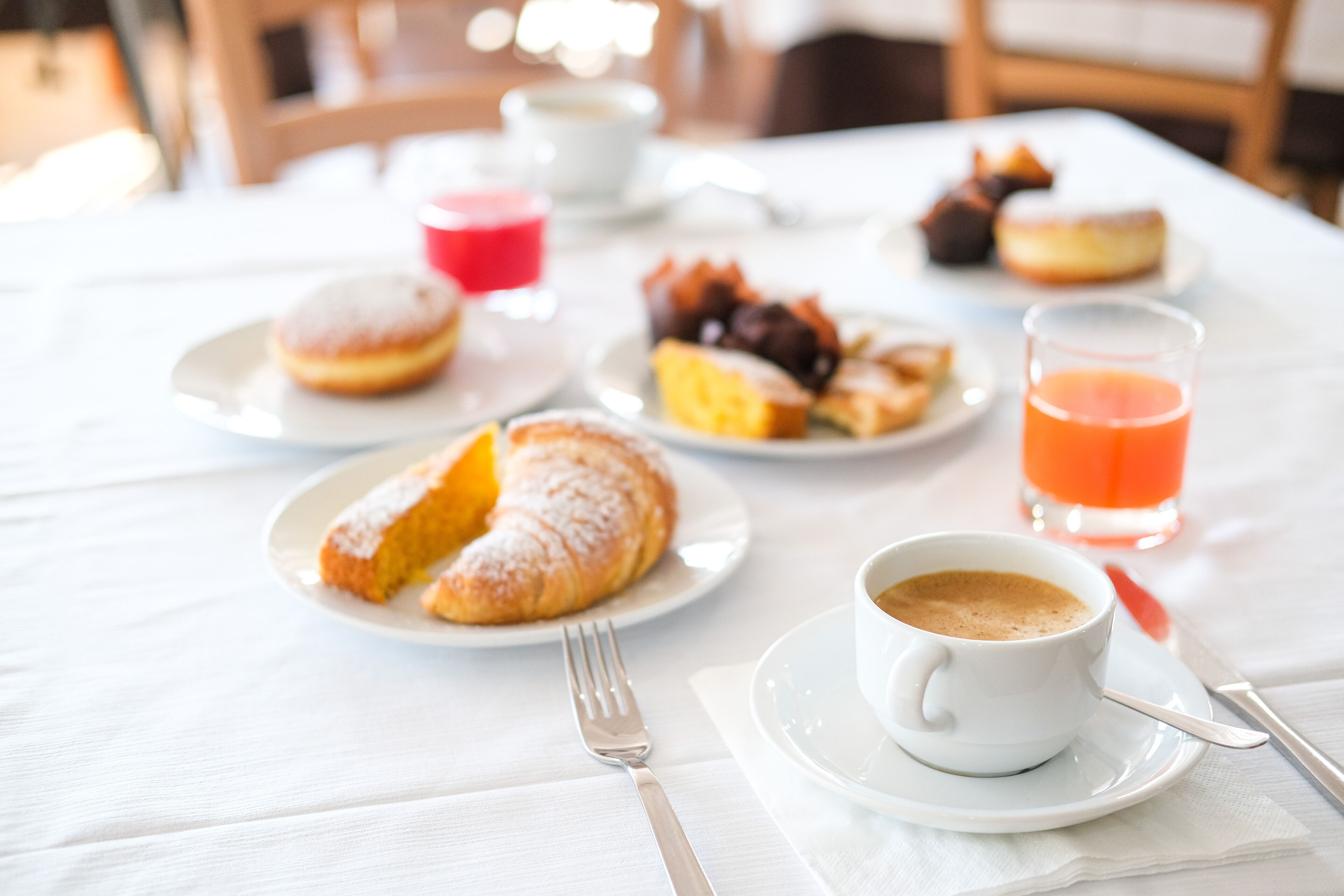 Mesa de café da manhã com doces, uma xícara de café e um copo de suco de laranja sobre uma toalha de mesa branca.