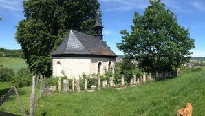 A small stone chapel with a pointed roof, surrounded by a stone fence and lush greenery.