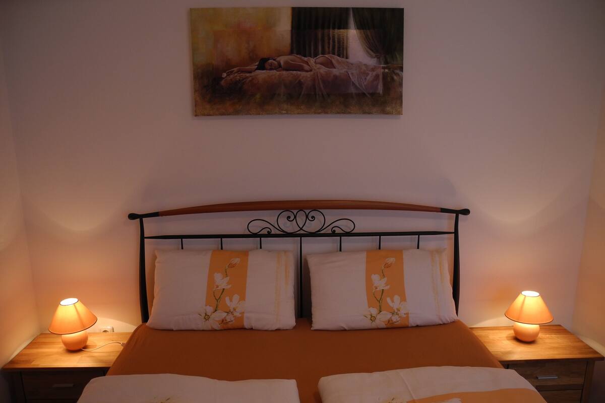 A neatly made bed with floral bedding, two bedside lamps, and a painting above the bed.
