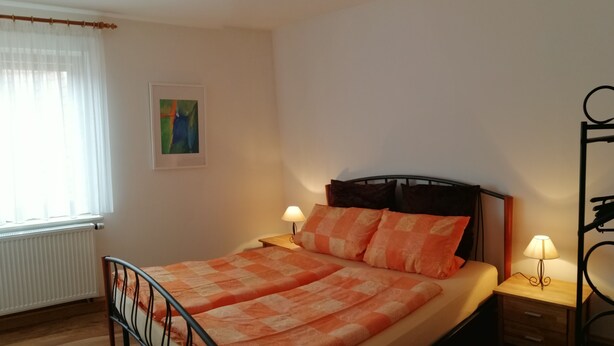 Un dormitorio con cama, dos mesitas de noche, un radiador, una ventana con cortinas y un cuadro en la pared.