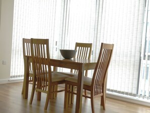 Enjoy meals in this dining area with a wooden table and four chairs, featuring a bowl on the table and large windows with vertical blinds.