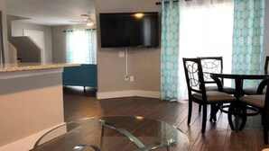 A modern living room with a glass coffee table, a TV mounted on the wall, and a dining area with chairs and a table. - Hamlet by I-4 · New! 5 Bedroom Pool Villa. Sleep 15+ Orlando (Kissimmee)