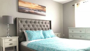 A bedroom with a tufted headboard, a bed with a floral patterned bedspread, a bedside table with a lamp, and a dresser. - Hamlet by I-4 · New! 5 Bedroom Pool Villa. Sleep 15+ Orlando (Kissimmee)