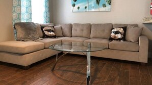 A living room with a beige sectional sofa, a glass coffee table, patterned curtains, and a painting of palm trees. - Hamlet by I-4 · New! 5 Bedroom Pool Villa. Sleep 15+ Orlando (Kissimmee)
