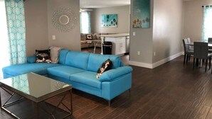 A living room with a blue sectional sofa, a glass coffee table, and a dining area with a table and chairs. - Hamlet by I-4 · New! 5 Bedroom Pool Villa. Sleep 15+ Orlando (Kissimmee)