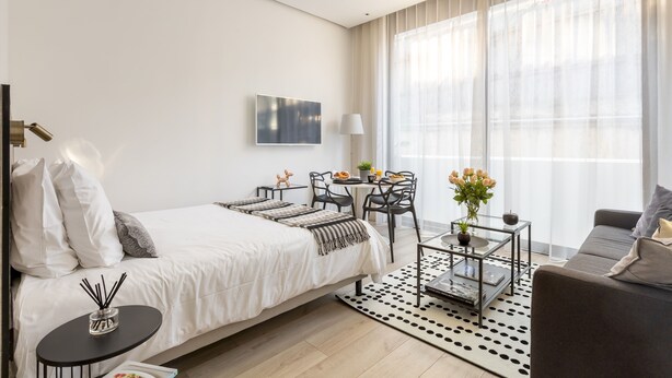 Una moderna camera d'albergo con un letto grande, un'area salotto con divano e tavolino, un tavolo da pranzo con sedie e un'ampia finestra con tende leggere.