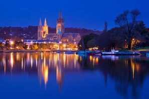 A historic building with a prominent clock tower, reflecting in a calm body of water, surrounded by trees and a dock with boats.