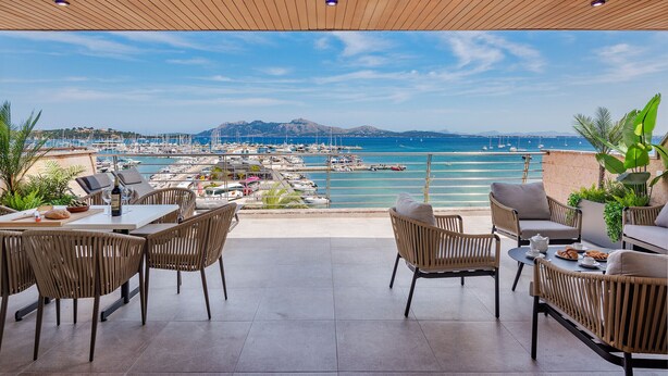 A terrace with wicker furniture, a table with a bottle, and a view of a marina with boats. - Vistamar IV frontline sea Apt. in a prime location in Puerto Pollensa (Puerto Pollensa)
