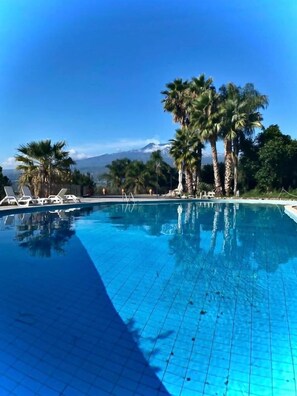 A swimming pool with clear blue water, surrounded by palm trees and lounge chairs. - Borgo San Biagio (Calatabiano)