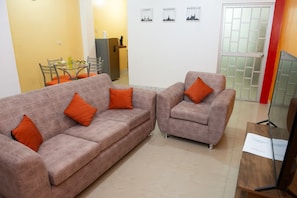 A living room with two sofas and a chair, a dining table with chairs, a refrigerator, and a TV on a wooden stand.